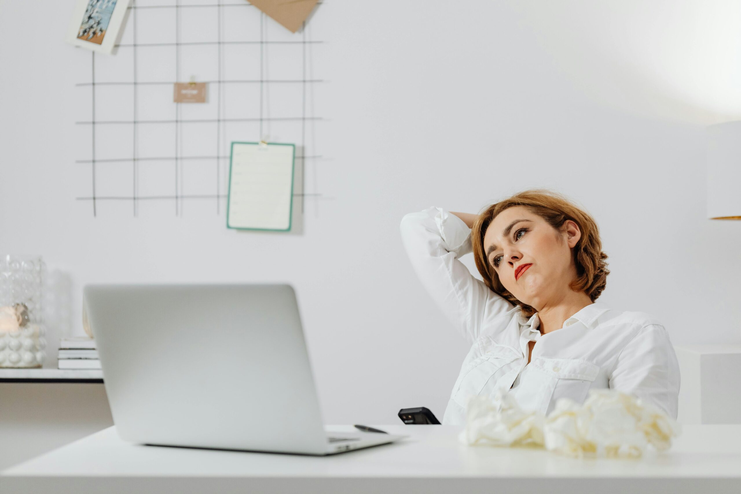 A woman in a white shirt sits at a desk with a laptop, appearing thoughtful indoors.