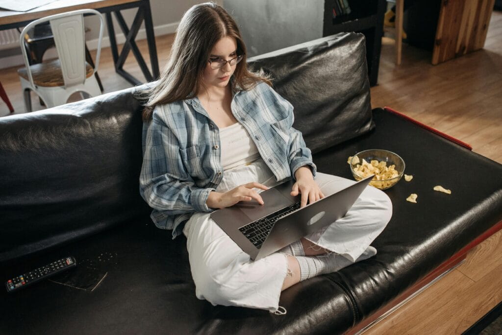 pexels-photo-7801204-7801204 Woman working on laptop in casual setting at home with snacks on sofa, depicting remote work lifestyle.